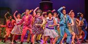 A colourful ensemble cast dances energetically on stage, with a central character wearing a sparkly dress and large hairstyle, surrounded by performer