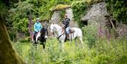 Two on horseback in green area next to old stone building