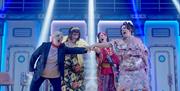 A scene from Menopause The Musical 2, featuring four actresses on stage in comedic poses. The set is designed like a cruise ship interior with colourf
