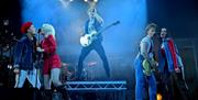 Energetic concert scene from 'Punk Off!' with a cast of performers. A lead guitarist takes centre stage on an elevated platform, surrounded by singers