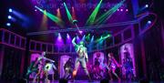 A dynamic ensemble cast performs on a colourful stage with vibrant green and purple lights. The central performer twirls a cane under a spotlight, sur