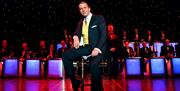 A stylish Stephen Triffitt as Frank Sinatra, seated centre stage in a classic suit and tie with a yellow waistcoat. The scene includes a live band in