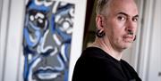 Artist Brian Farrell stands in front of one of his abstract portraits, arms folded and facing the camera. He has grey hair and facial piercings, inclu