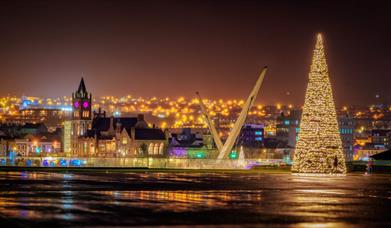 Derry at Christmas - picture of Ebrington Square Christmas Tree with Peace Bridge and Guildhall in the background.