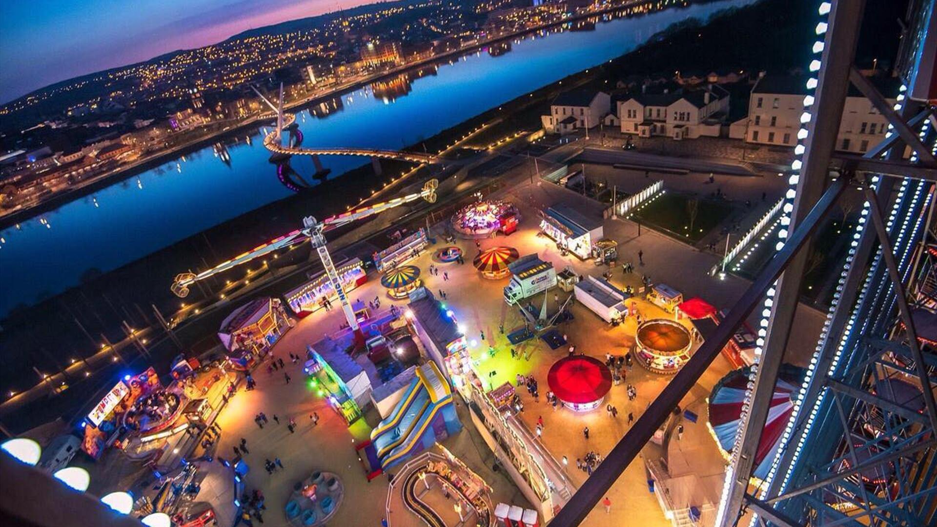 Birds eye view of a funfair beside a river at night-time.