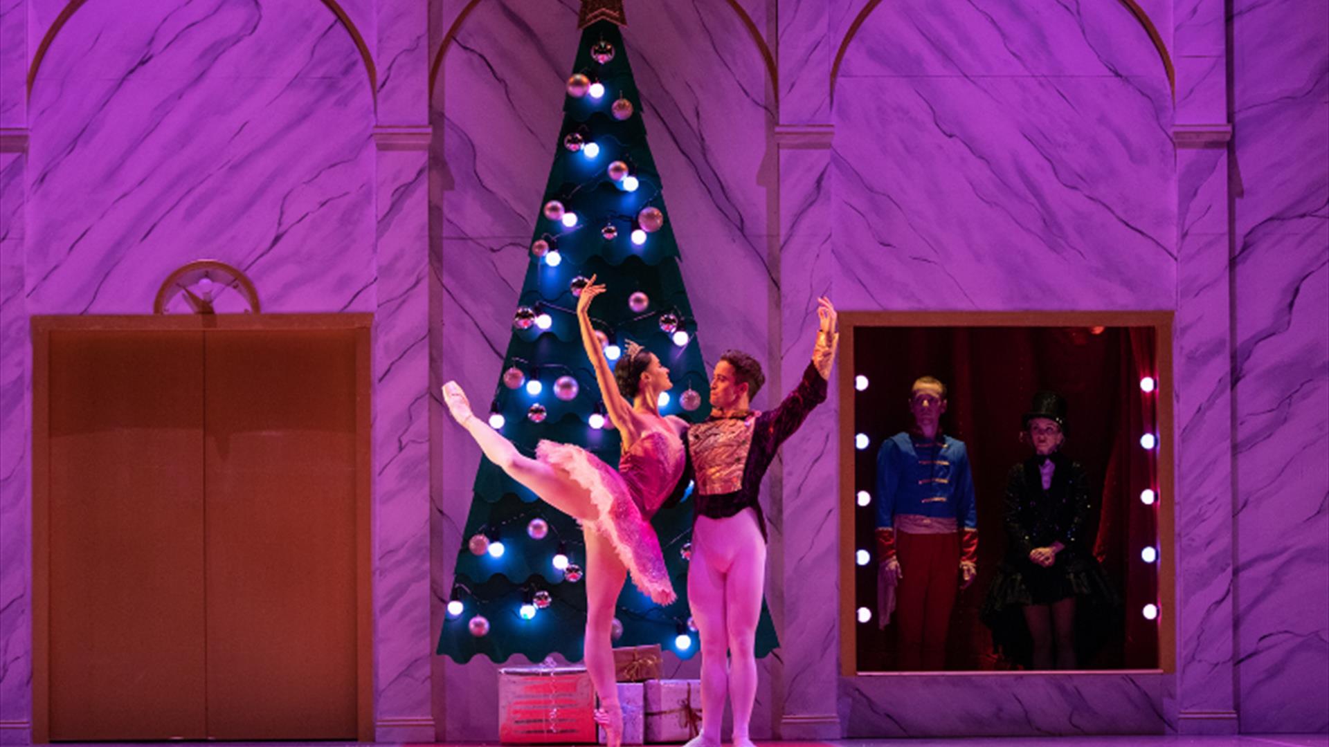 Ballerina Clara in arabesque and The Nutcracker, in front of a sparkling Christmas tree, arms extended in pose. Soft purple lighting