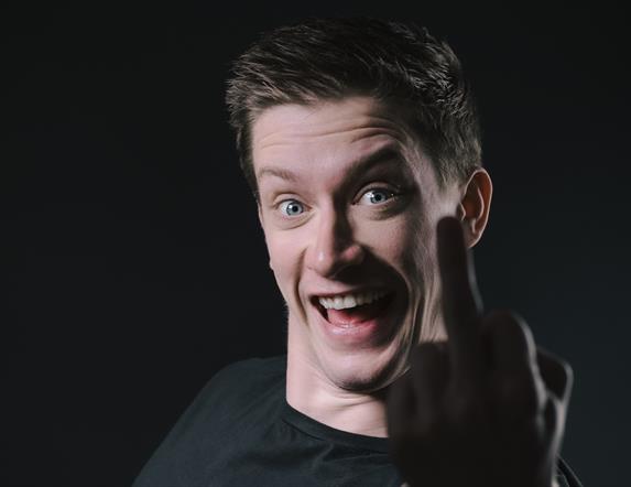 Daniel Sloss, grinning in an animated fashion, with the silhouette of his middle finger in front of his face. Against an all-black background.