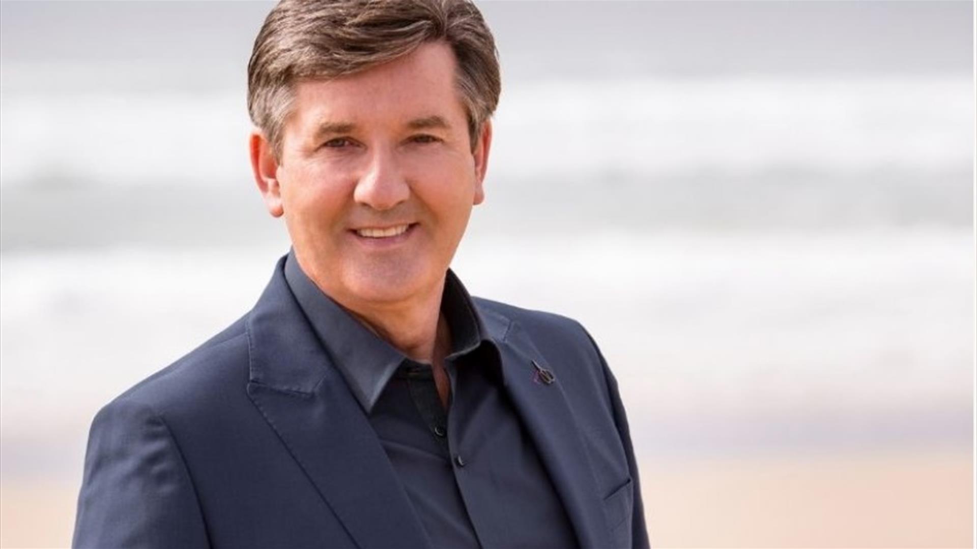 Daniel O'Donnell pictured in a black suit and shirt, smiling in front of a blurred background