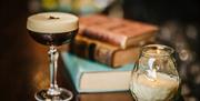 an Espresso Martini cocktail situated beside some old books and a candle in a glass jar