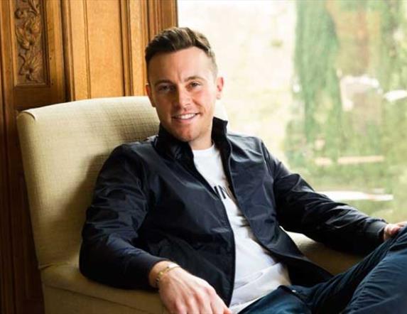 Nathan Carter, sitting and smiling leisurely in an armchair, in front of a window  with a view of greenery.