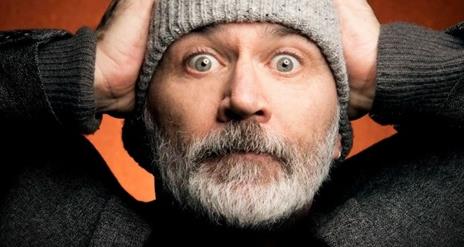 Tommy Tiernan wearing a grey hat, with his hands n his head- looking mildly exasperated against a burnt orange background.