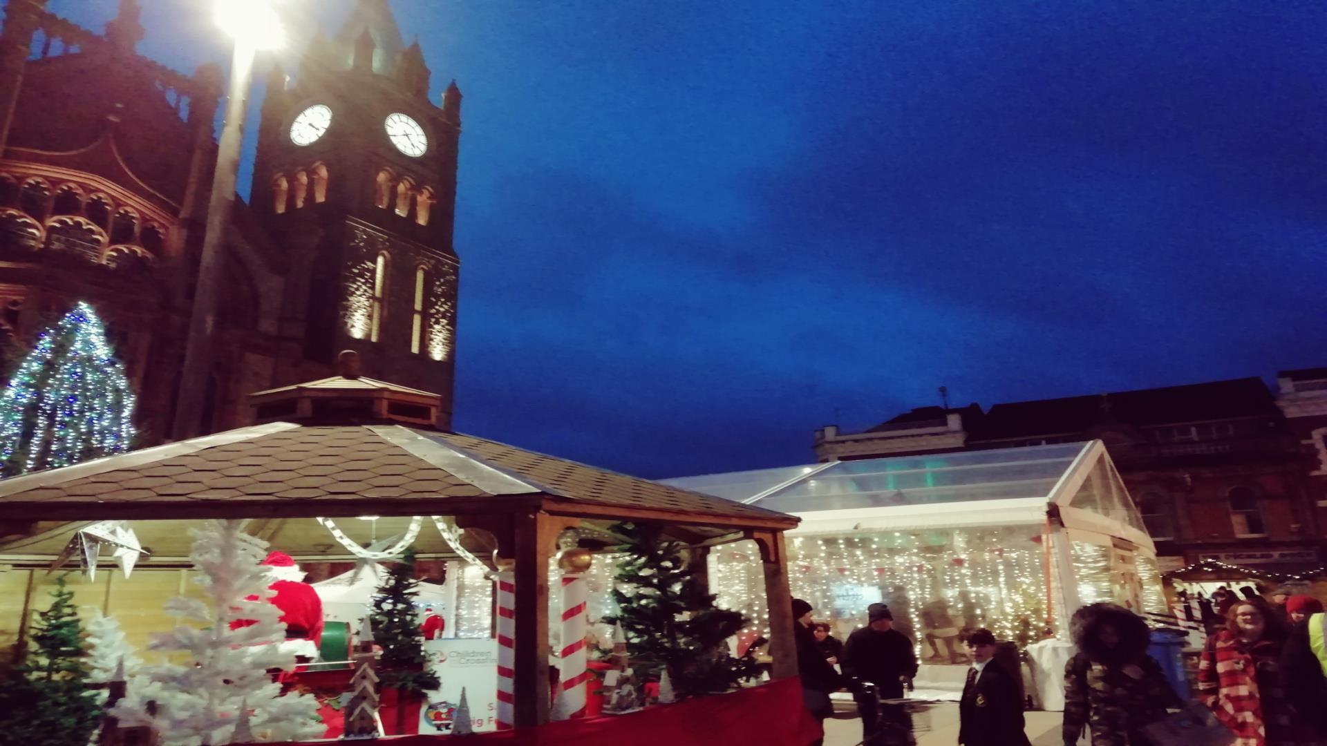image of the Walled City Markets at Christmas at night. Christmas stalls with Guidhall and Guildhall clock in the background clo