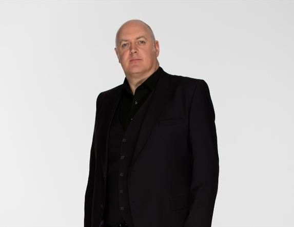 Dara O'Briain, sporting a neutral expression, standing in the centre in all black, against a white background