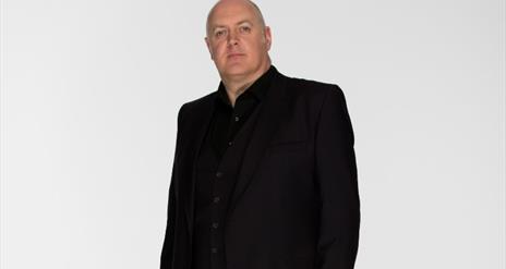 Dara O'Briain, sporting a neutral expression, standing in the centre in all black, against a white background