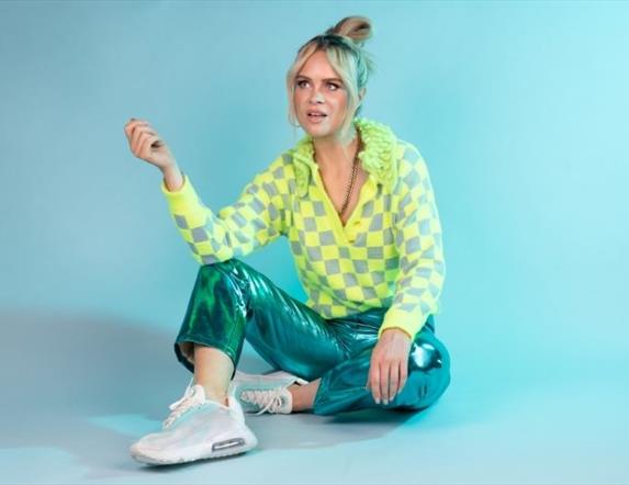 Joanne sitting casually on the floor in a bright yellow and turquoise outfit, against a cool green/blue background.