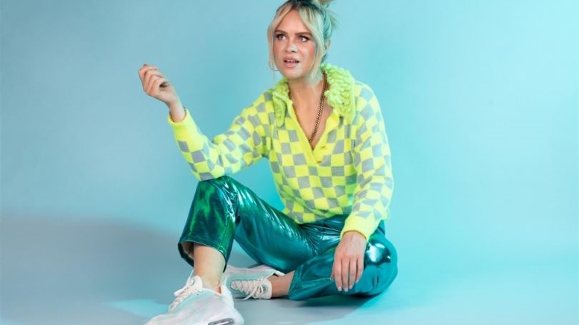 Joanne sitting casually on the floor in a bright yellow and turquoise outfit, against a cool green/blue background.