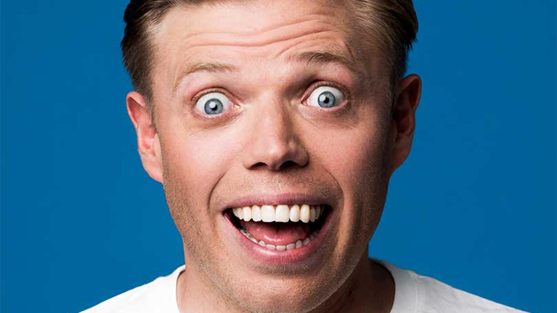 Rob Beckett Smiling widely in front of a deep blue background