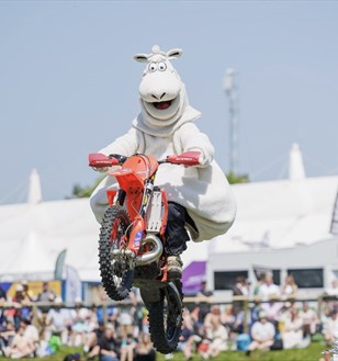 A person in a sheep costume performs a motorcycle jump at an outdoor event, with a crowd watching in the background. The scene is lively and entertain