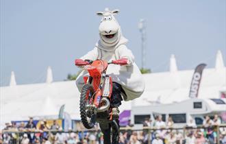 A person in a sheep costume performs a motorcycle jump at an outdoor event, with a crowd watching in the background. The scene is lively and entertain
