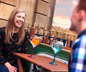 woman and man sat outdoors smiling whilst drinking a cocktail 