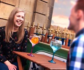 woman and man sat outdoors smiling whilst drinking a cocktail 