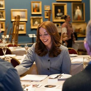 Photograph of a woman leading a discussion at a dining table inside an art gallery, surrounded by framed paintings and seated attendees. Warm lighting