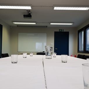 Large white table with drinking glasses and a water jug.  Flipchart, whiteboard, two doors, one window