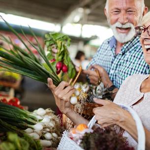 Woman and man smiling next to vegetable stall of market. Woman holding vegetables.