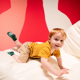 Photograph of a young child with red hair wearing a mustard yellow shirt and beige shorts, smiling while crawling on a large white cushioned surface. 