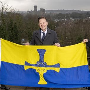 The County Durham Flag being held by 3 people. Durham Cathedral in background.