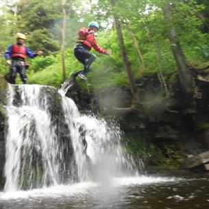 2 children in life vests and safety helmets jumping from some rocks into a river with a small waterfall below them