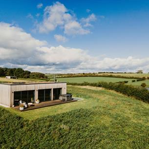 Modern wooden lodge with large glass windows and hot tub, set in a grassy hilltop overlooking green fields and a pond under a blue sky with scattered 