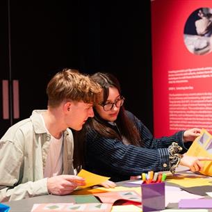 A young man with brown hair and a young woman with brown hair and glasses sat together making art in a bright pink space.