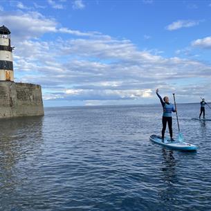 Two people paddleboarding near a weathered lighthouse on a calm sea under a blue sky with scattered clouds.