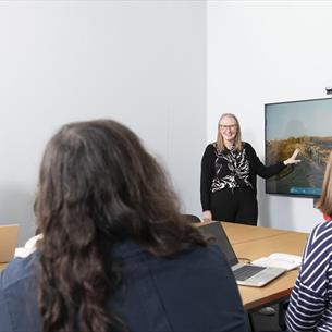 Meeting room with 4 people, three seated at table and one standing by display screen.