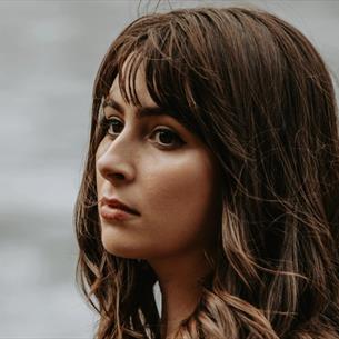 A women with brown hair looking out into the distance, there's a blurred grey background behind her.