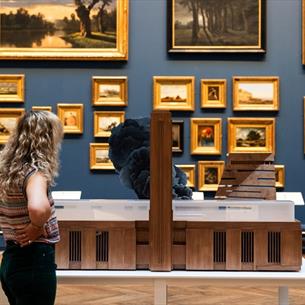 A woman with blonde curly hair looking at Tate Modern on Fire by Nathan Coley in a blue picture gallery with walls covered by paintings