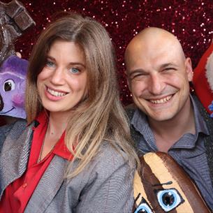 Two performers smiling at the camera surrounded by colourful puppets.