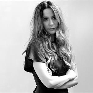 Black and white photograph of a woman with long wavy hair standing with arms crossed, facing slightly to the side. Background is plain with a draped f