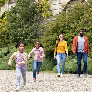 A family, 2 adults and 2 children, running in the grounds of The Bowes Museum, smiling.