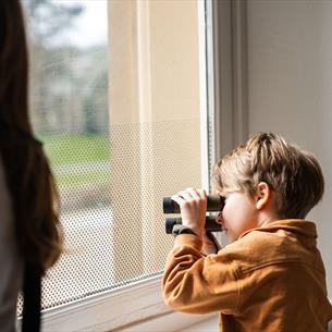 A young boy looking through binoculars out of a window, with a blurred figure of a woman in the foreground. 