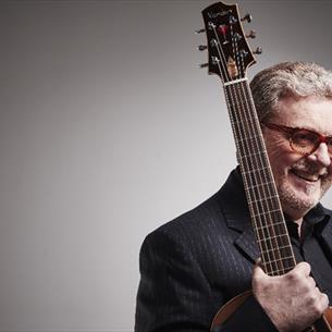 A man holding an acoustic guitar vertically against his shoulder, set in a studio with two bright lights positioned behind him on either