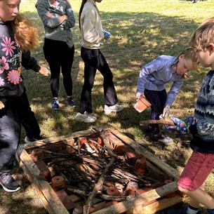 A group of children gathered around a wooden frame filled with sticks and bricks on grass, engaged in a hands-on outdoor activity. 