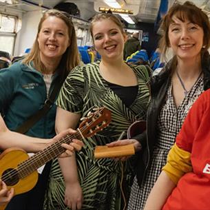 Sam Slatcher smiling and playing guitar on public transport, standing with four women who are also smiling. 