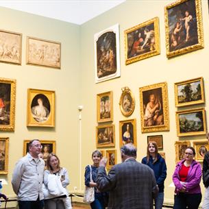 A group of ten people listening attentively to a curator in a museum gallery filled with framed historical paintings.