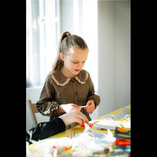 A young girl in a brown dress with hair in a ponytail, holding something made of paper, with craft materials on a yellow table in front 