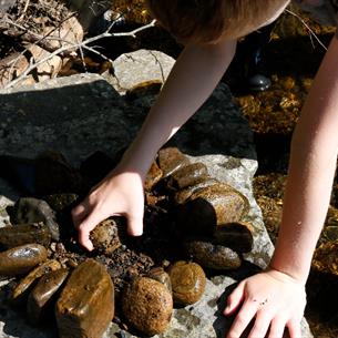 A child in rubber boots leans over a rock by a clear stream, arranging small stones into a circular pattern