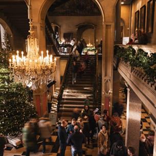Photograph of a grand interior space with festive decoration, featuring a large Christmas tree with lights and ornaments, a chandelier, and garlands.