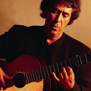 A man playing a classical guitar, seated against a plain, warm-toned background. The man wears a dark suit, focusing on his fingers.