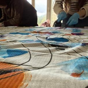 Two people working on a large fabric with colourful abstract patterns in blue, orange, and black lines. Focus is on fabric surface.